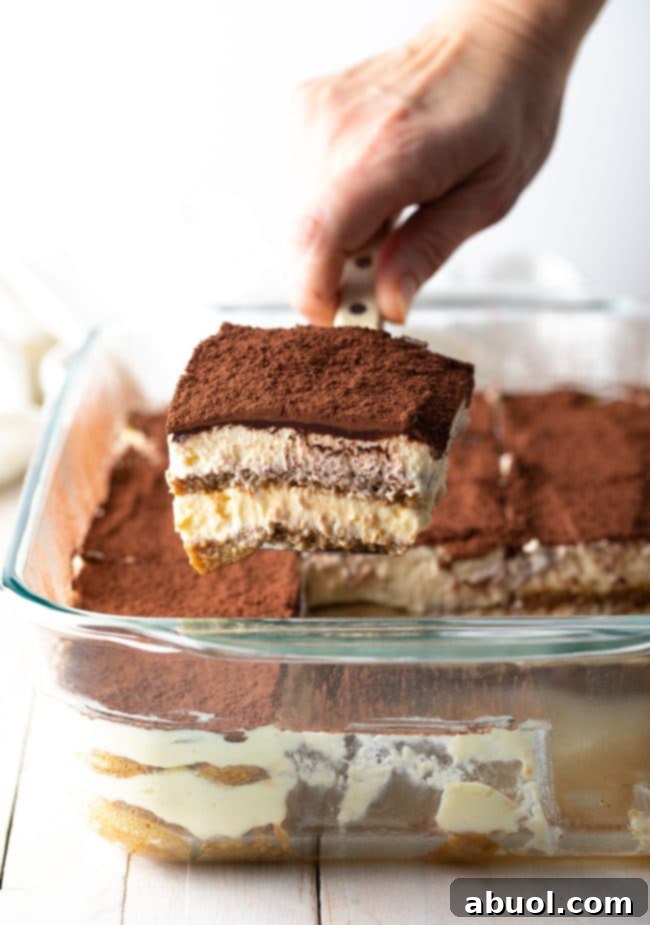 A hand carefully holds a spatula presenting a perfect piece of easy tiramisu, with the remaining cake visible in the background, showcasing its inviting layers.
