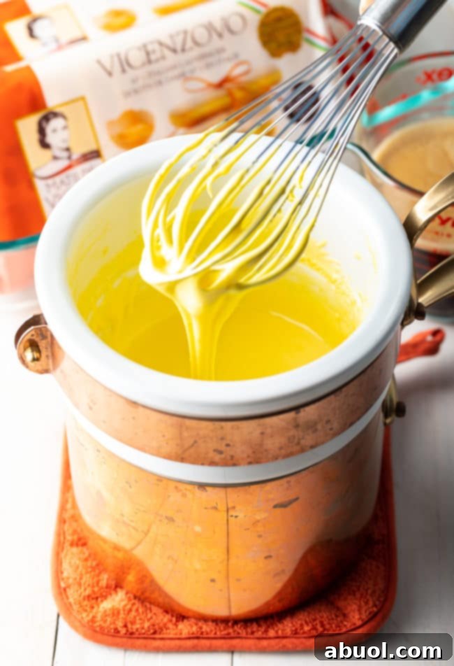 A whisk vigorously mixes the sabayon mixture in a glass bowl set over simmering water, creating a light and airy base.