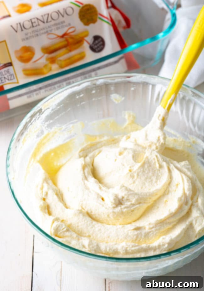 A rubber spatula gently combines the creamy mascarpone and freshly whipped heavy cream in a spacious glass bowl, creating the signature Tiramisu filling.