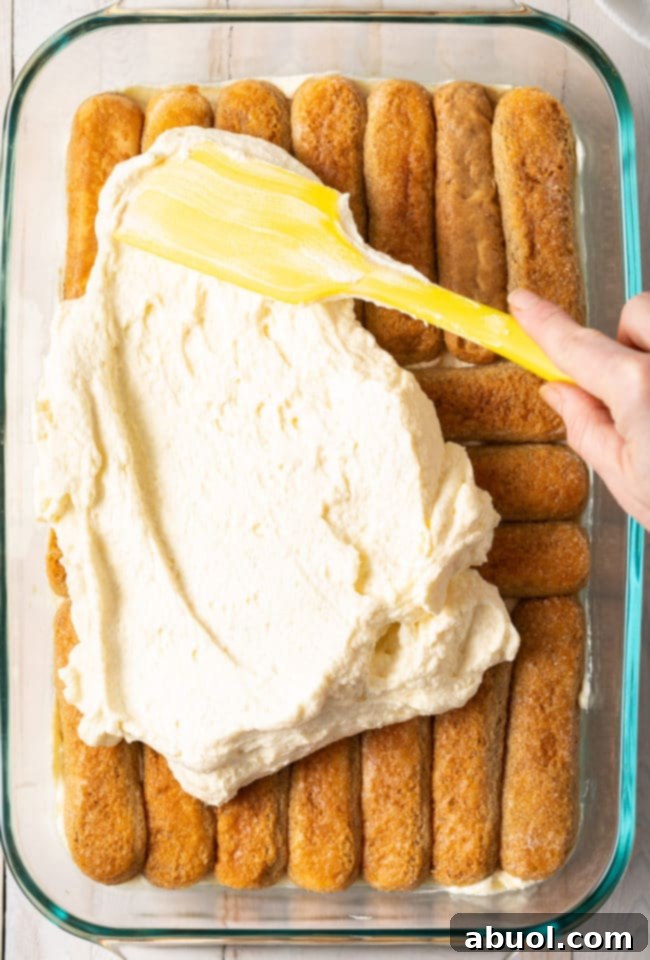 A spatula smoothly spreads the rich mascarpone mixture evenly over the layer of coffee-soaked ladyfingers, ensuring full coverage and creamy indulgence.