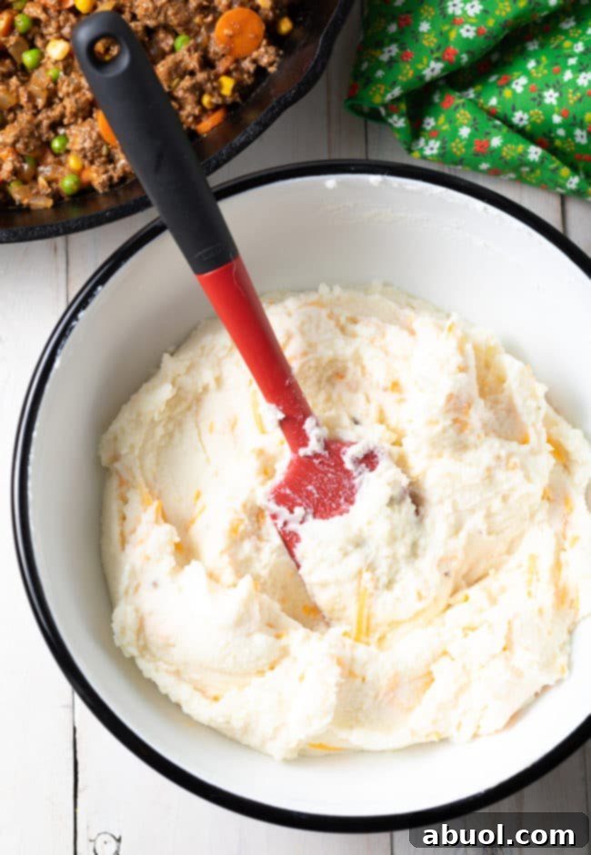 Creamy mashed potatoes being generously mixed with shredded cheddar cheese in a large mixing bowl, preparing for the Cottage Pie topping.