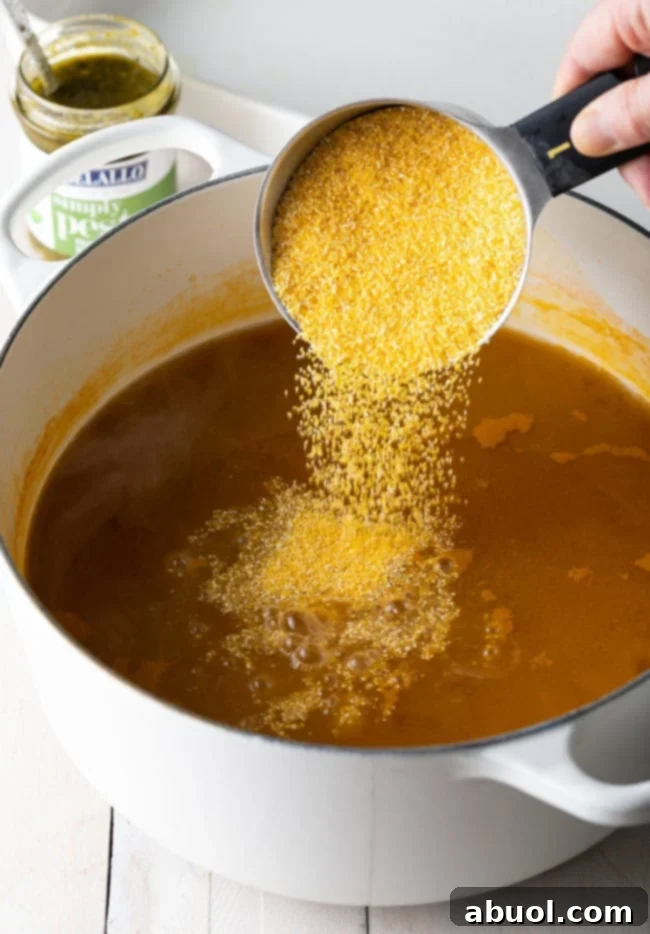 Hand adding dry polenta from a measuring cup into a large pot of simmering broth.