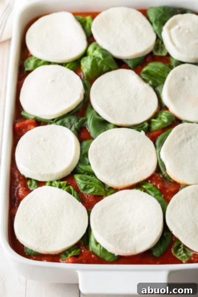 White baking dish with layers of marinara sauce, fresh basil leaves, and slices of mozzarella cheese.