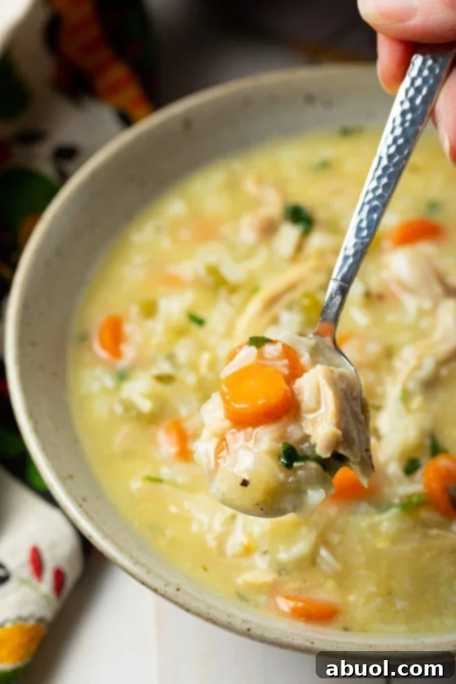 A hand holds a spoon filled with creamy chicken and rice soup, glistening over a white bowl, ready to be enjoyed. Fresh parsley garnish adds a touch of color.