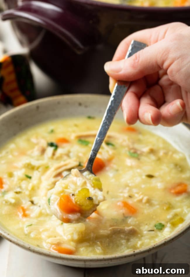 A hand holds a spoon with a generous scoop of creamy chicken and rice soup over a white bowl, ready to be eaten. A sprinkle of fresh green herbs adds a finishing touch.