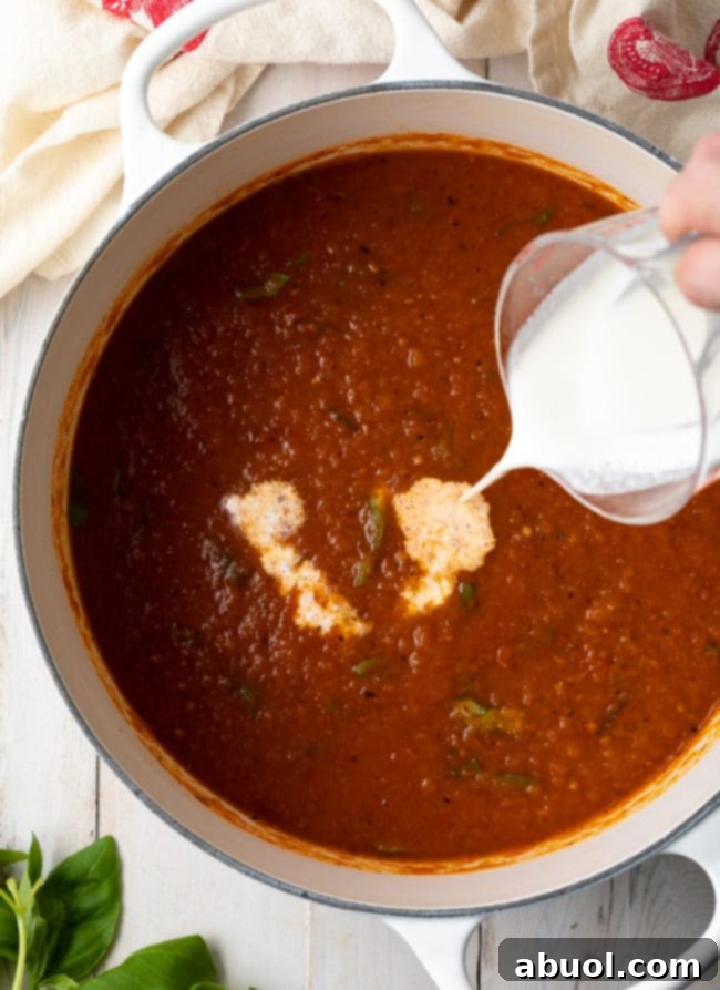 Roasted Tomato and Basil Velvet 9 Tomato soup in a pot with a hand pouring heavy cream into the soup.