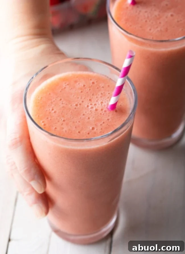 A hand holding a glass of strawberry banana smoothie with a straw.