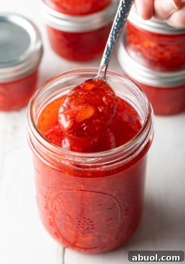 A hand holding a spoonful of vibrant homemade strawberry freezer jam above a rustic glass jar, illustrating the perfect consistency and rich color of a freshly made batch.