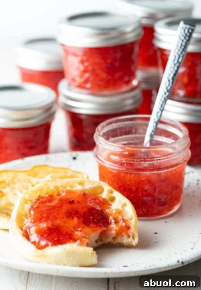 An English muffin with a bite taken out, generously spread with homemade strawberry freezer jam, paired with a matching jar of jam, highlighting the appeal of this fresh preserve.