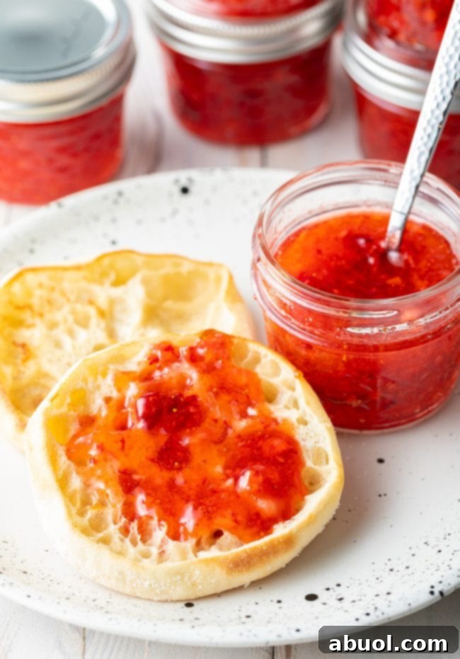 A perfectly toasted English muffin generously topped with homemade strawberry freezer jam, alongside a matching jar of jam, showcasing a delightful breakfast or snack.