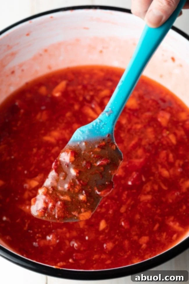 A hand holding a blue spatula, showing off a perfectly textured dollop of freshly made strawberry freezer jam, ready for jarring.