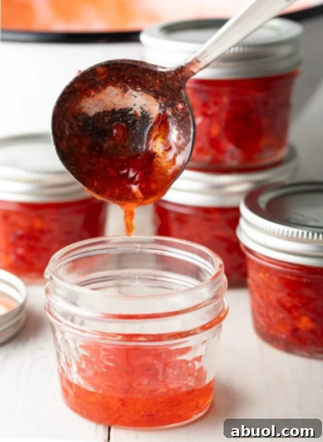 A close-up of vibrant strawberry jam being carefully ladled into a clear glass canning jar, ready for setting and freezing.