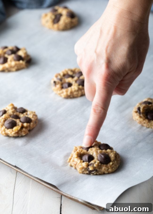 A hand gently points to a perfectly melted chocolate chip embedded in a warm, freshly baked banana oat cookie on parchment paper.
