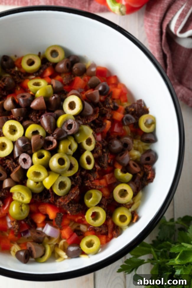 Close-up of olives, cooked orzo, sun-dried tomatoes, and diced red peppers in a mixing bowl, ready to be combined.