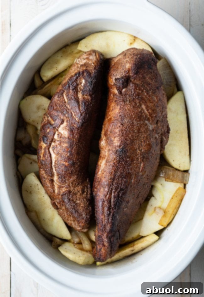 A perfectly seasoned pork tenderloin, rich with a spice rub, resting atop a generous bed of fresh apples and sautéed onions in the crock pot, ready for slow cooking.