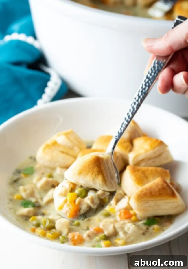 A hand holding a spoon with a perfect bite of creamy chicken pot pie mixture, topped with a piece of biscuit.