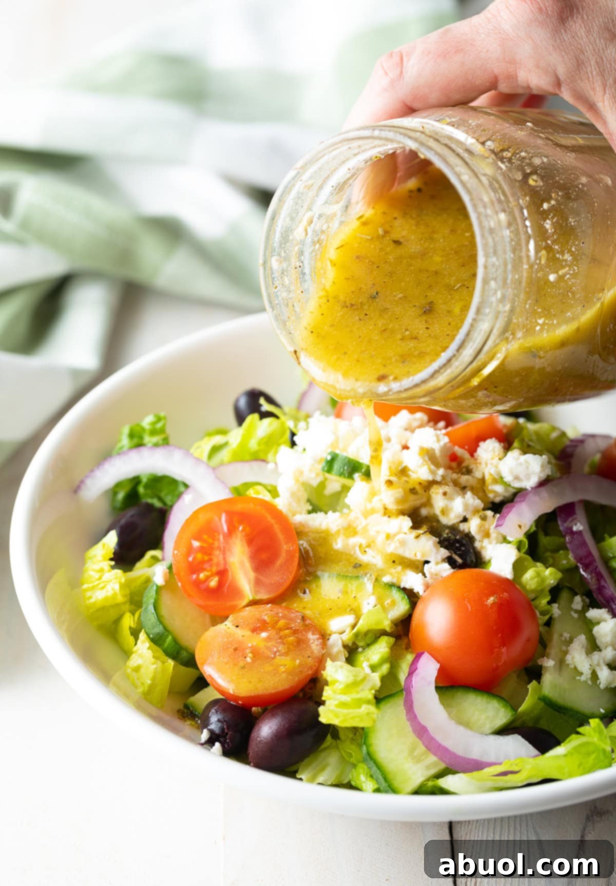 Zesty Homemade Greek Dressing 9 A hand pouring a glass of homemade Greek Salad Dressing over a vibrant Greek Salad, ready to be enjoyed.