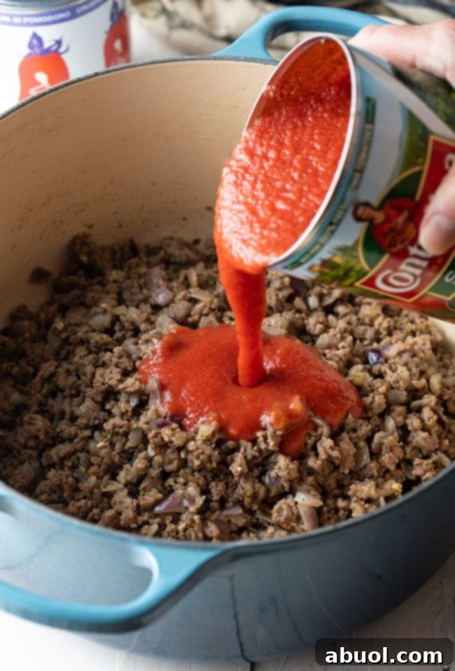 Hand pouring canned tomato sauce into a pot with browned ground beef and other ingredients