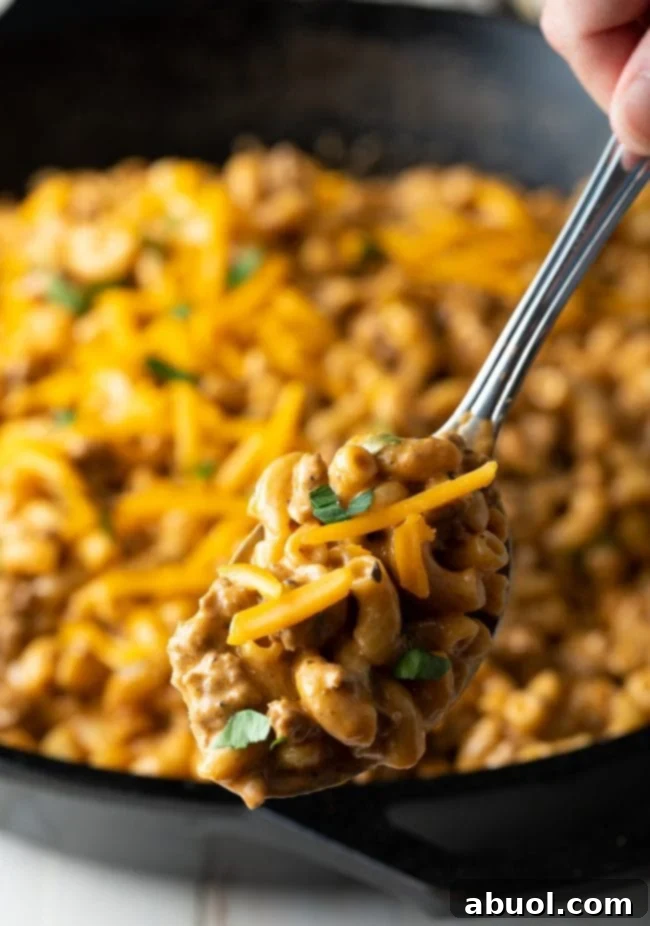 A metal spoon lifts a generous serving of creamy cheeseburger macaroni from a skillet.