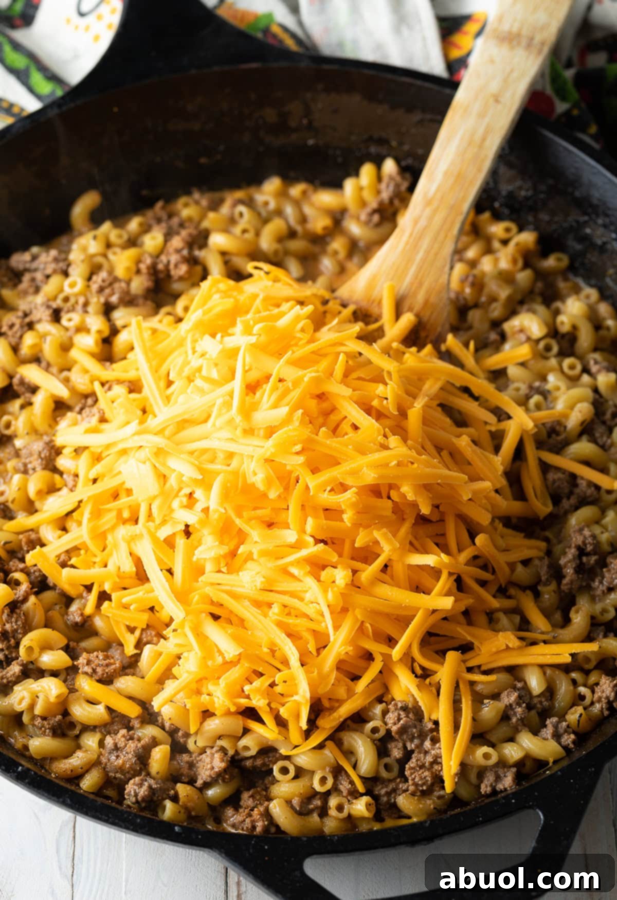 A hand stirring shredded cheddar cheese into the beef and macaroni mixture in a skillet.