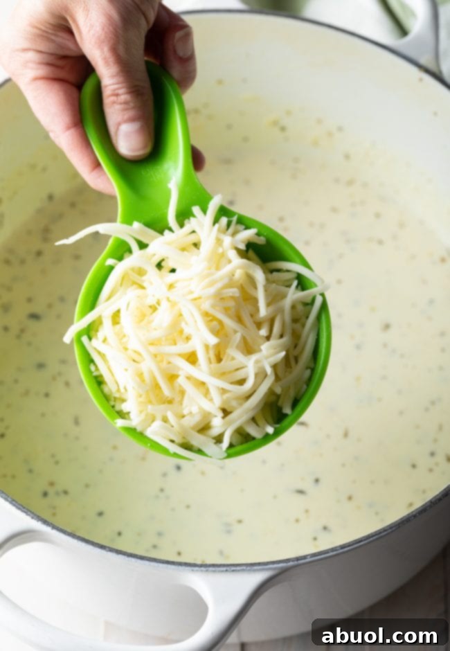Hand holding a green measuring cup filled with freshly shredded mozzarella cheese, ready to be added to the dish.