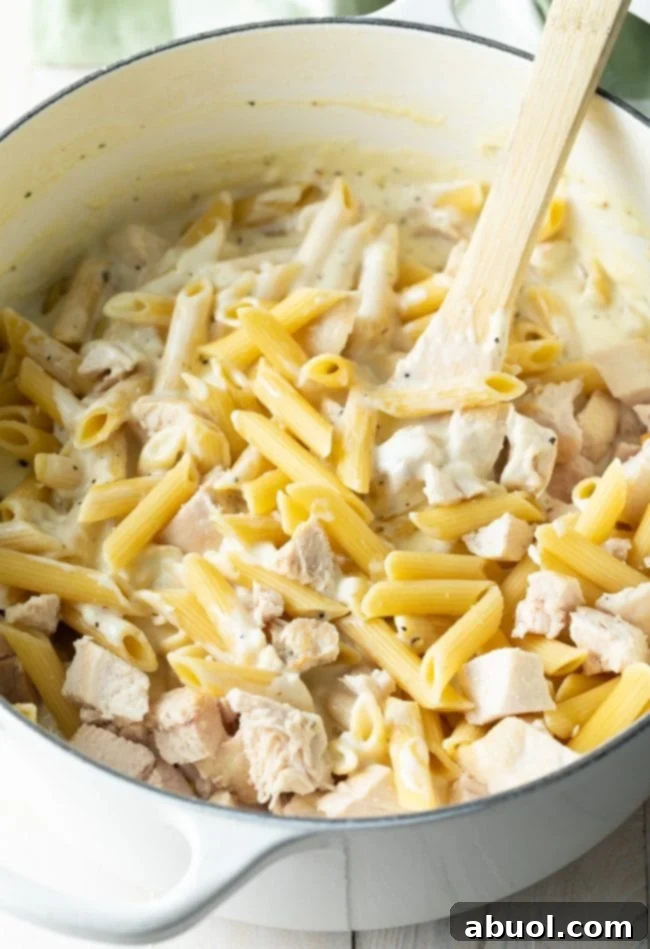 Large cooking pot showing penne pasta, cooked chicken chunks, and creamy Alfredo sauce being stirred with a wooden spoon.