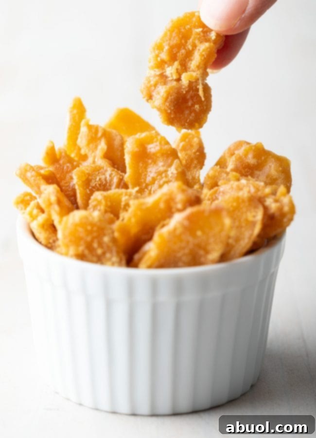 A hand gently taking a piece of golden homemade crystallized ginger candy from a small ceramic ramekin, with more candies in the background.