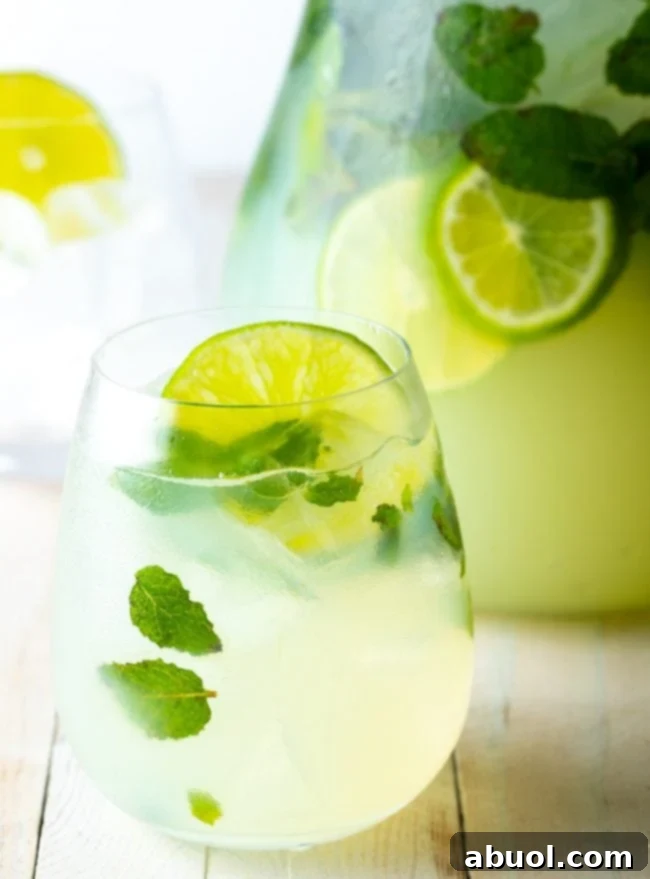 A refreshing glass of mint mojito with a pitcher of the drink in the background, garnished with fresh lime and mint sprigs.