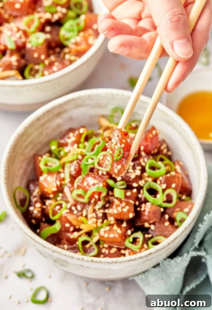 A hand holds a piece of raw tuna with chopsticks, with a bowl of vibrant Ahi Poke blurred in the background, highlighting the freshness of the ingredients.
