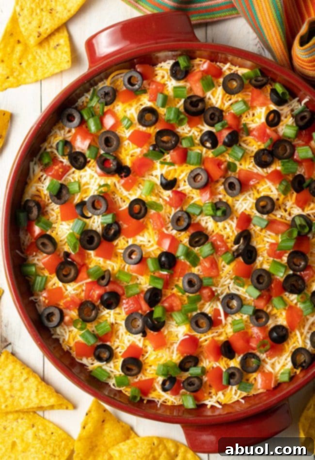 Top down view of a vibrant red round dish brimming with Layered Mexican Taco Pizza Dip. The surface is beautifully adorned with melted shredded cheese, vibrant chopped green onions, perfectly sliced black olives, and juicy diced tomatoes. The dish is invitingly surrounded by a generous scattering of crispy tortilla chips, ready for dipping.