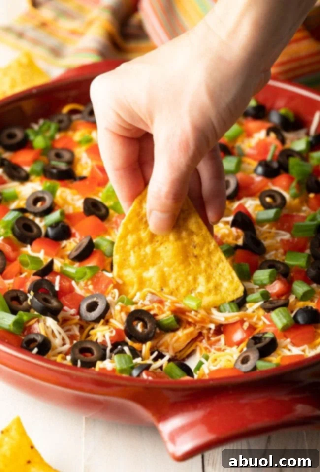 A hand expertly scoops into the creamy, layered Mexican taco pizza dip using a crispy tortilla chip. The dip showcases a delectable blend of shredded cheddar cheese, thinly sliced green onions, rich black olives, and juicy chopped tomatoes, all ready to be savored.