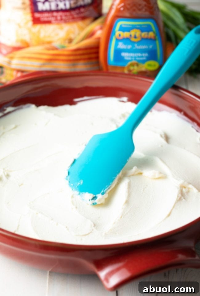 A vibrant red pizza pan is being prepared with a smooth layer of cream cheese, expertly spread using a blue spatula, forming the foundational base for the Layered Taco Pizza Dip.