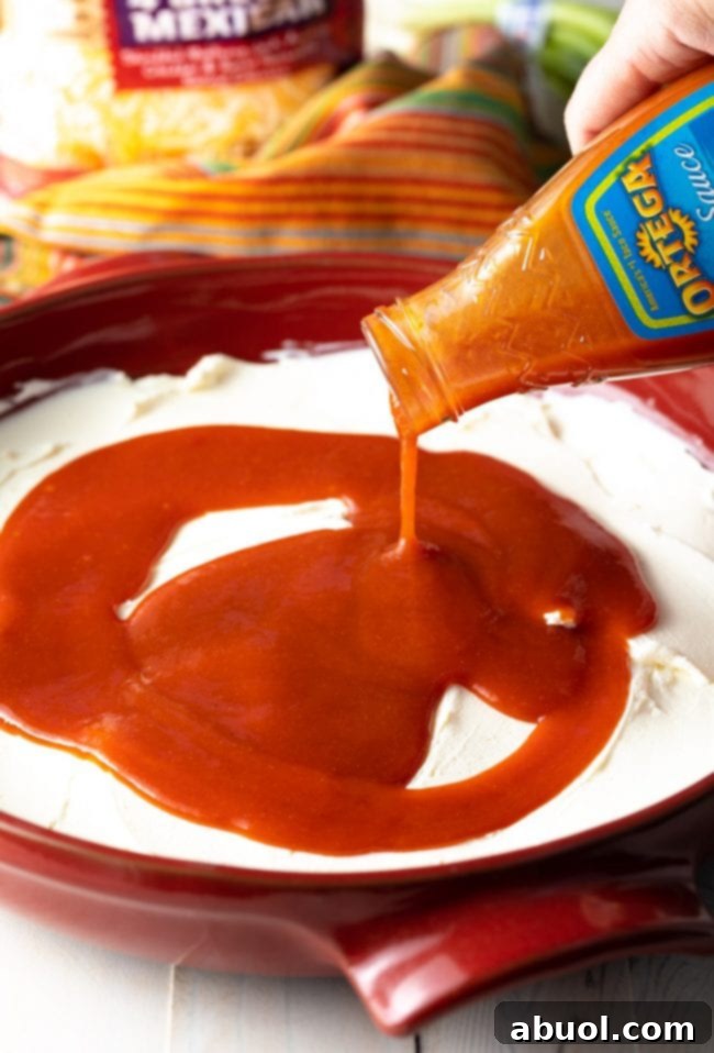 A stream of vibrant enchilada salsa is being carefully poured over a smooth layer of cream cheese spread in a red round baking dish, creating the next delicious layer of the dip.