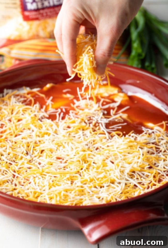 A hand is gently sprinkling a generous amount of shredded cheese over the freshly spread enchilada sauce, adding a delicious and visually appealing layer to the dip.
