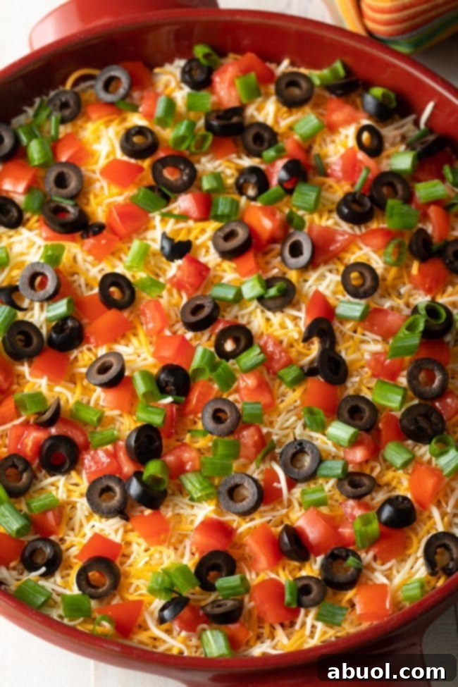 A captivating top-down view of the freshly prepared Layered Taco Pizza Dip, elegantly presented in a round dish. The surface is adorned with a rich scattering of shredded cheddar cheese, vibrant sliced green onions, distinct black olives, and juicy chopped tomatoes, showcasing its tempting layers and colorful appeal.