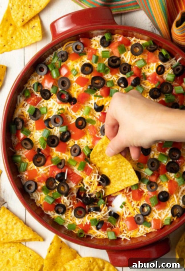 A hand gently scoops a generous portion of Mexican pizza dip with a tortilla chip. The dip is beautifully adorned with shredded cheddar cheese, vibrant green onions, savory black olives, and fresh diced tomatoes, showcasing its readiness to be enjoyed.