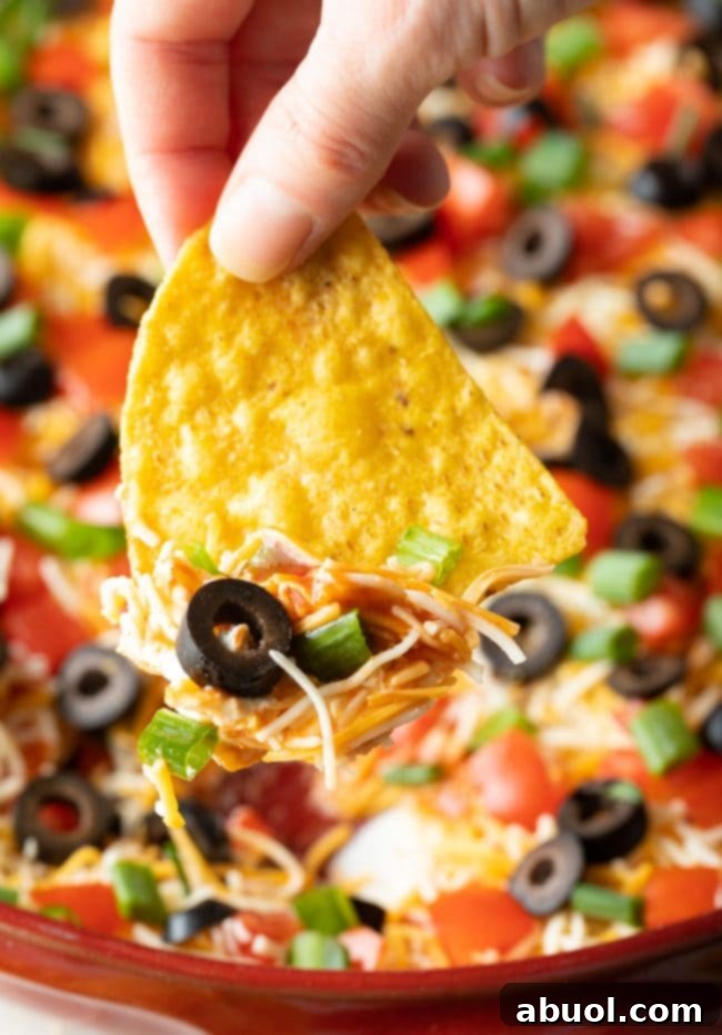 A hand reaches into a layered Mexican pizza dip with a tortilla chip, capturing a scoop of shredded cheddar cheese, sliced green onions, black olives, and diced tomatoes, highlighting the vibrant and irresistible layers of the appetizer.