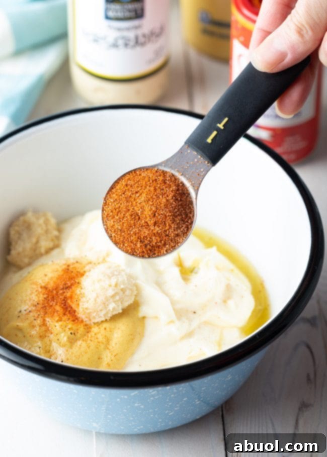 Hand holding a measuring spoon full of Cajun seasoning over a bowl with the rest of the ingredients. 