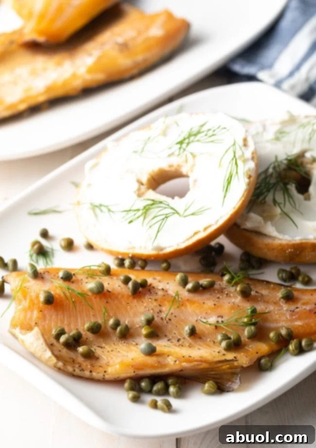 Smoked trout fillet on a white plate topped with capers next to a bagel. 