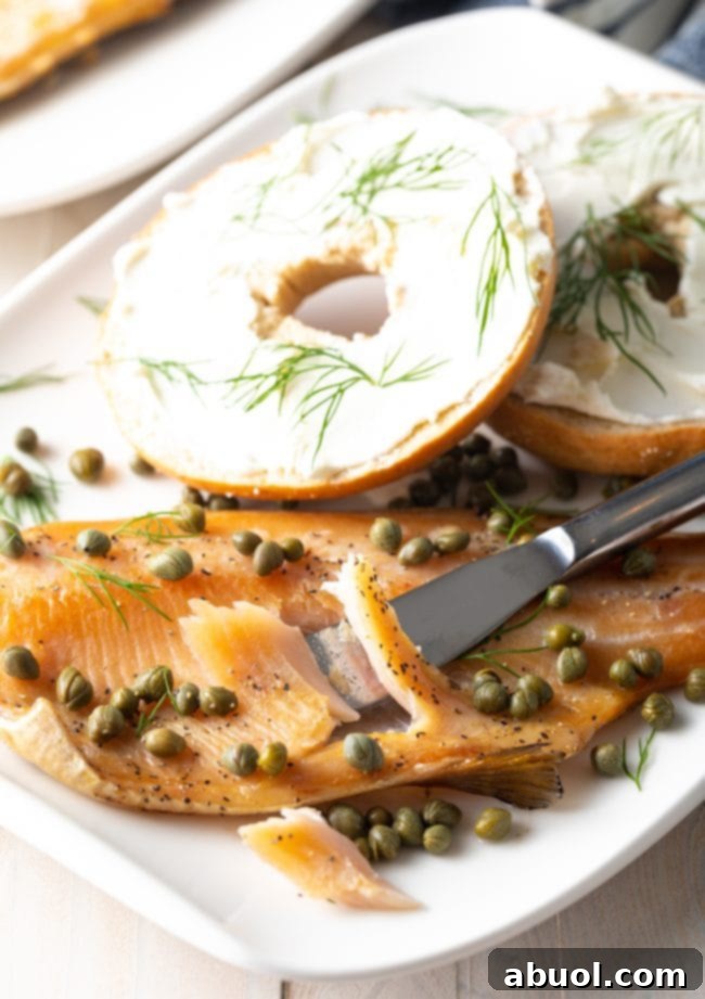 Flaky smoked trout being flaked apart with a knife on a plate next to a bagel. 