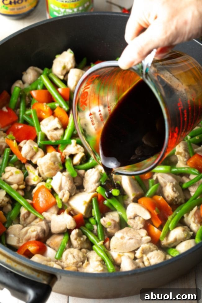 The savory sauce being poured over the stir-fried chicken and vegetables in the pan, mixing to coat all ingredients.