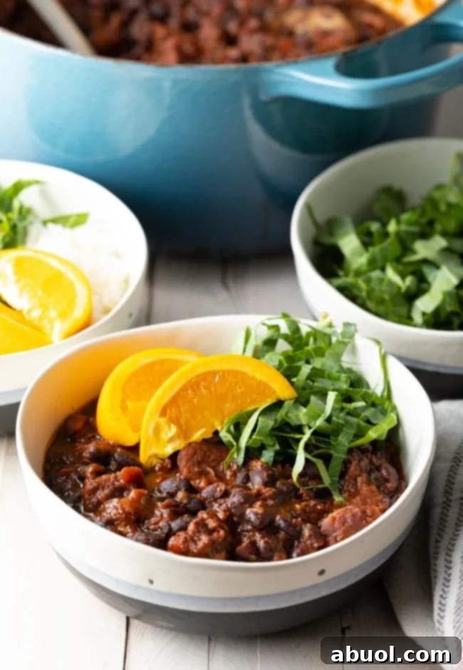 A steaming bowl of traditional Brazilian Feijoada, a pork and black bean stew, garnished with bright orange slices and finely shredded green vegetables. In the softly blurred background, additional bowls of fresh garnishes and the large cooking pot suggest a communal, inviting meal.