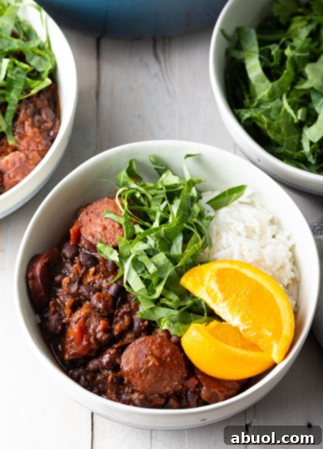 A bowl of Brazilian Feijoada, a hearty pork and black bean stew, is artfully topped with vibrant orange slices and finely shredded collard greens. In the soft focus background, additional garnishes and the large cooking pot hint at a festive and abundant meal setting.