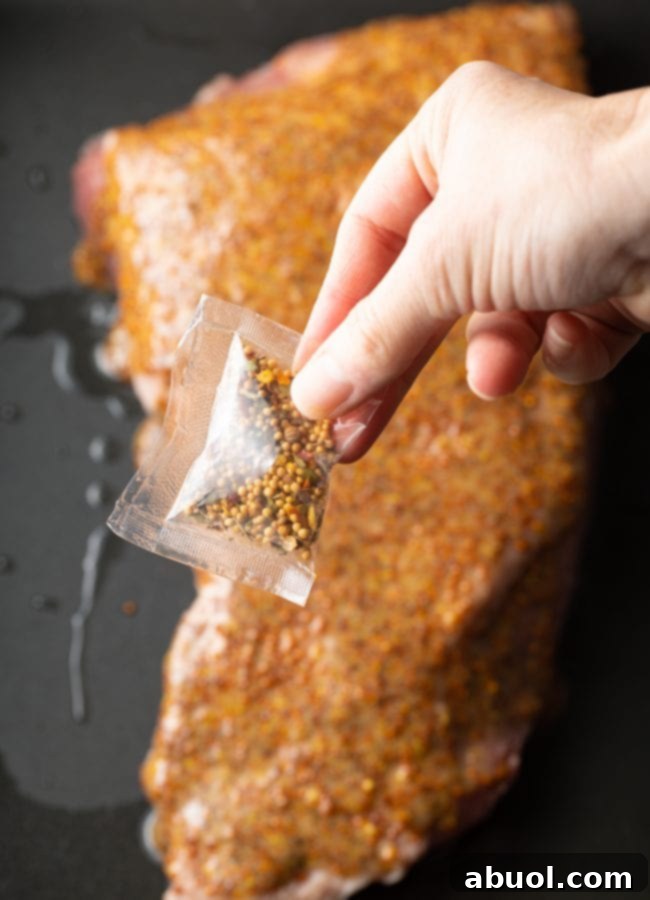 A hand holding the spice packet that comes with corned beef, poised above the raw brisket in a roasting pan.