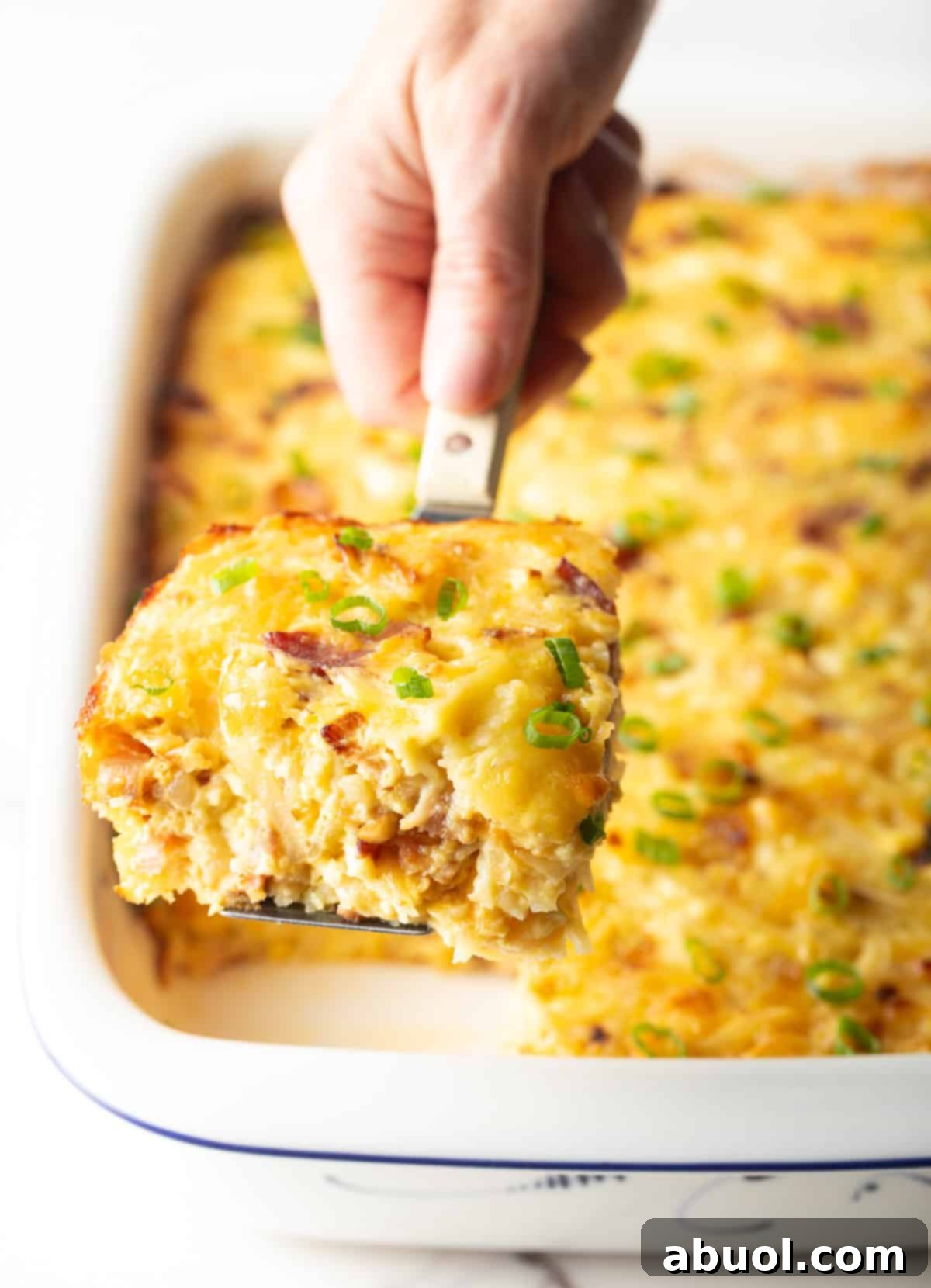 Amish Breakfast Casserole: Hand using a metal spatula to lift a square of Amish breakfast casserole from a white baking dish.