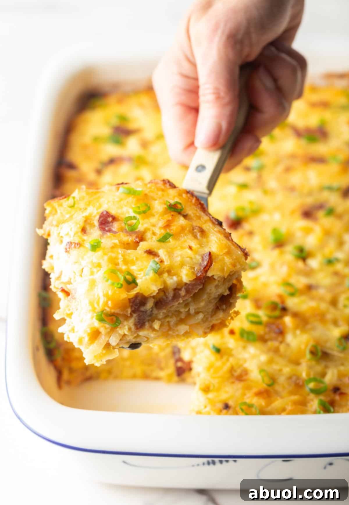 Amish Breakfast Casserole: A hand using a metal spatula to lift a square of Amish breakfast casserole from a white baking dish.