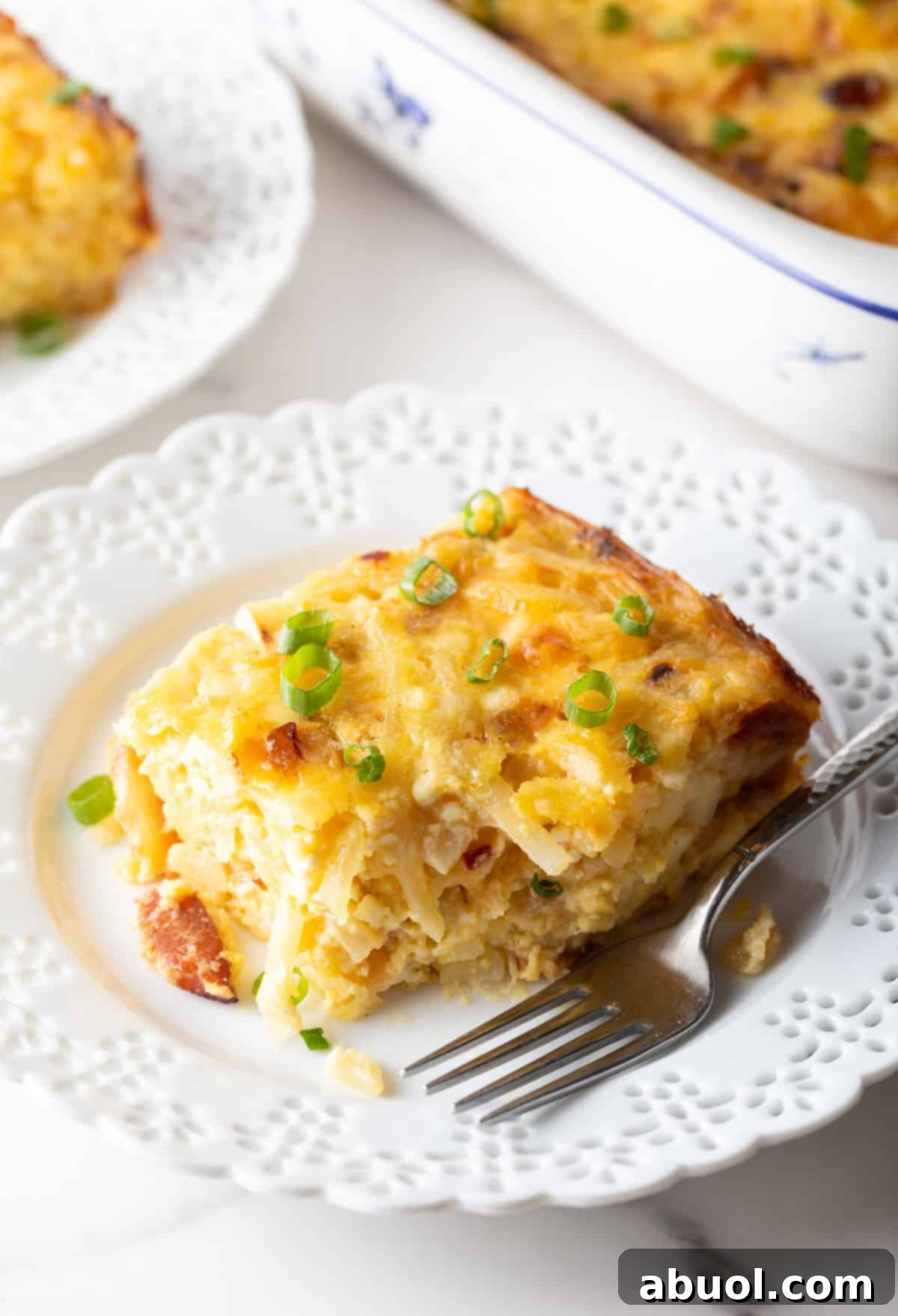 Amish Breakfast Casserole: Square serving of hash brown potato, egg and cheese casserole on a white plate with a fork.