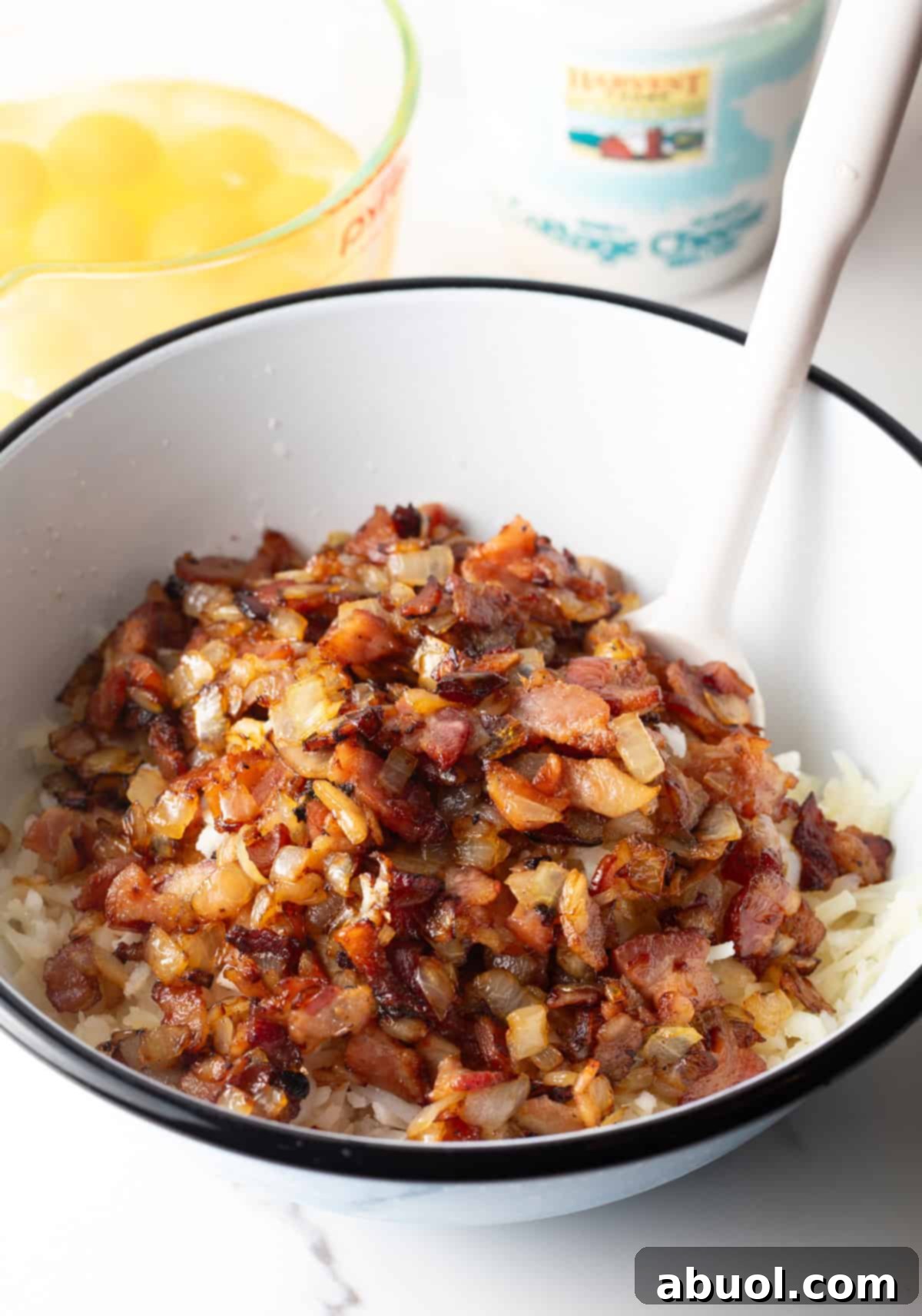 Amish Breakfast Casserole: Adding cooked chopped bacon to a white mixing bowl with shredded potatoes.