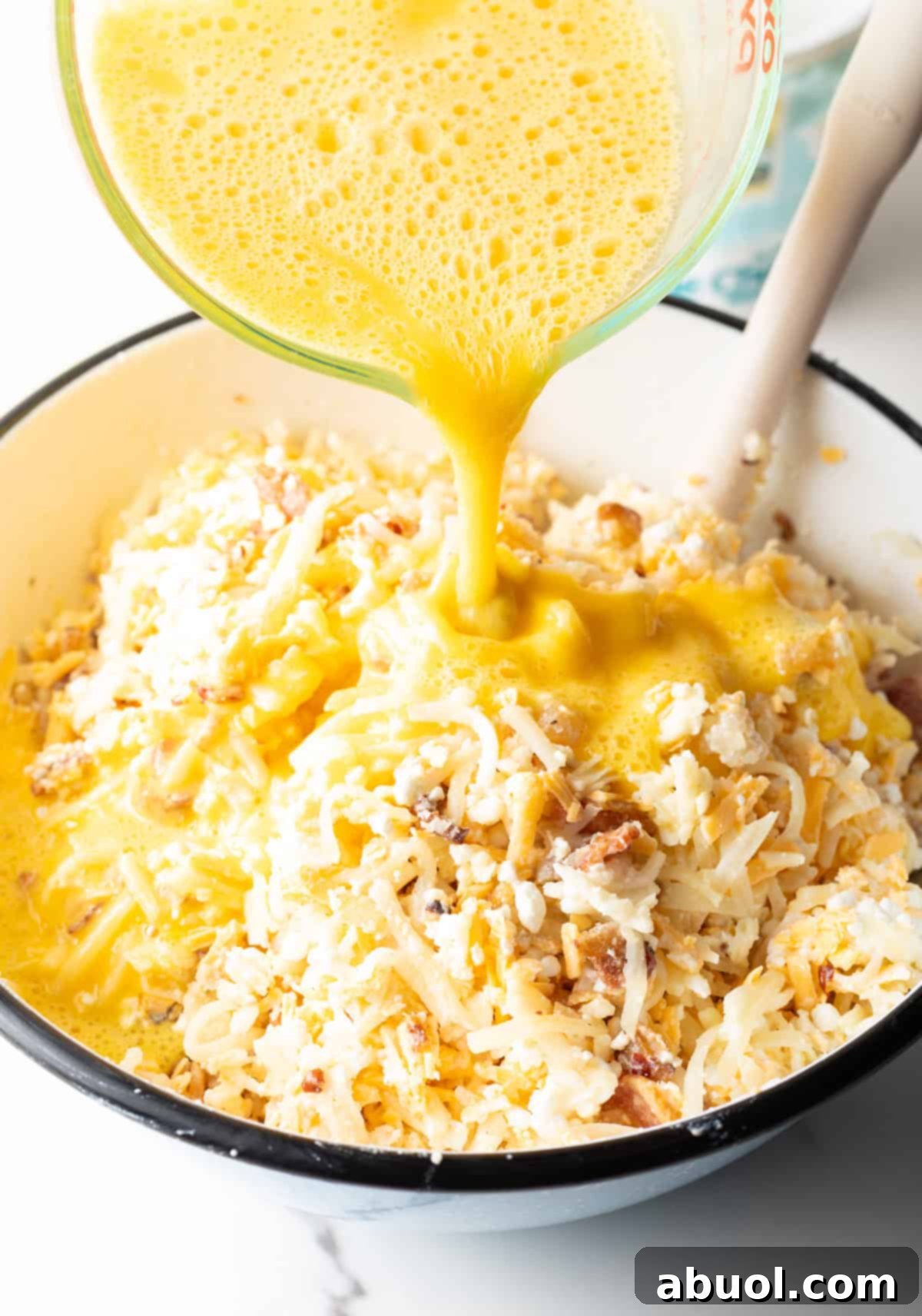 Amish Breakfast Casserole: Pouring egg mixture from a glass measuring pitcher into a white bowl with thawed hash brown potatoes, cheese and bacon.