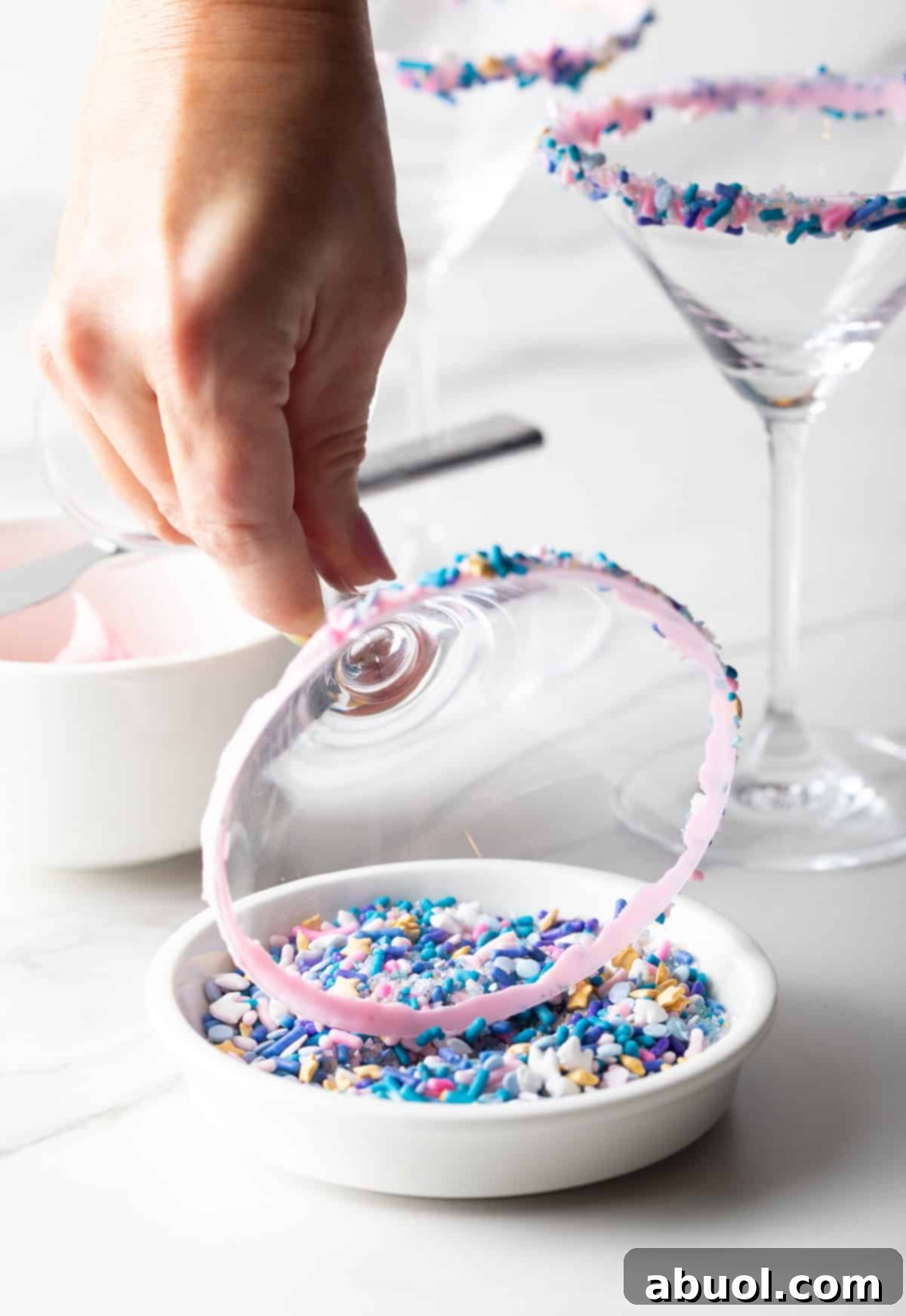 A martini glass being dipped into colorful sprinkles to line the rim, illustrating the fun and easy process of creating the signature Sugar Cookie Martini garnish.